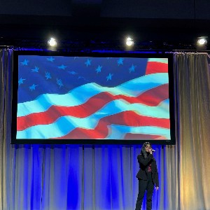 Shamini Sathya Prakash singing the National Anthem with an American flag projected behind her
